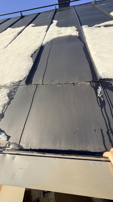 Metalltak med smältande snö vid listerna och skarv. Möjliga läckage kan ses där solen ligger på och snön smälter.