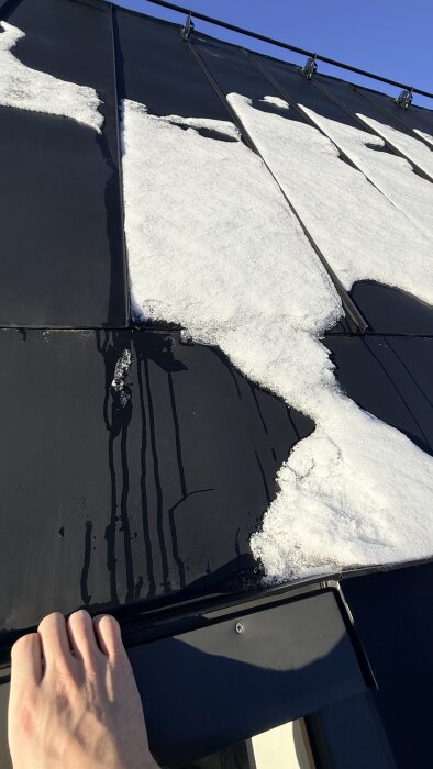 En hand håller i kanten av ett tak med snöfläckar smältande på svart takplåt under en klarblå himmel.