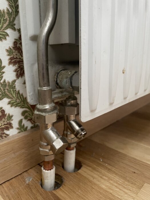 Radiatoranslutning med vita rör och metallventiler på ett trägolv framför en blommig tapet.