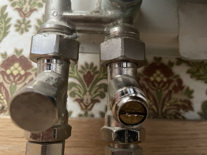Två metallrörkopplingar under en diskbänk med mönstrad tapet i bakgrunden.