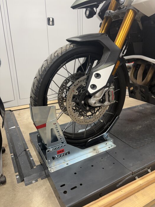 Framhjul på motorcykel placerat i ett fäste på en lyftplattform i ett garage, omgiven av förvaringsskåp och verktyg.