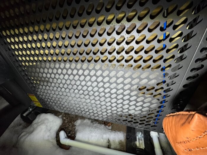 Utomhusbild på perforerat metallgaller med snö vid basen och del av orange klädesplagg synligt i hörnet. Ett vitt avloppsrör ses nedtill.