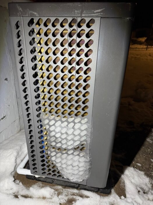 Utedel av en luftvärmepump med isbildning på gallret, omgiven av snö.
