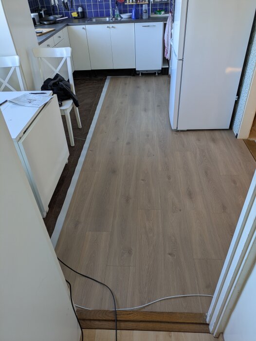 Kök under renovering med nylagt trägolv, synlig kyl och köksbänk med lila kakel i bakgrunden.