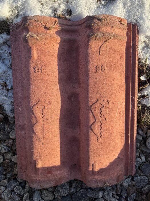 Röd betongpanna av modell Zanda Nordica från 90-talet, liggande på grus och snö. Märkta med nummer 96 och lätt väderpåverkade.