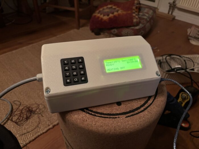 Vit styrbox med LCD-skärm och knappsats, visar aktuell och inställd temperatur samt status för uppvärmning. Boxen står på korkunderlag i ett rum.