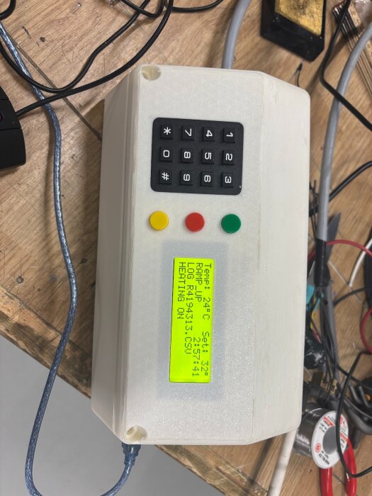 En vit låda med en knappsats och skärm, ansluten till kablar på ett träbord, används som en del av ett projekt för styrning av en keramikugn.