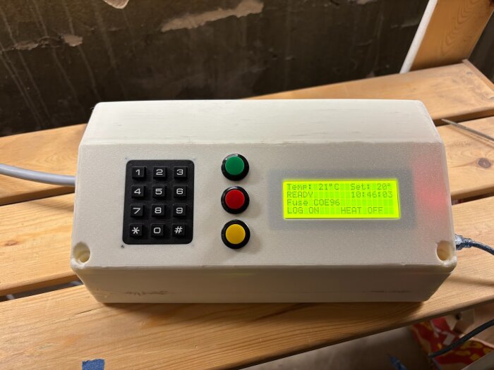 Kontrollbox med knappsats och display som visar temperaturinställningar och status, placerad på en trähylla.