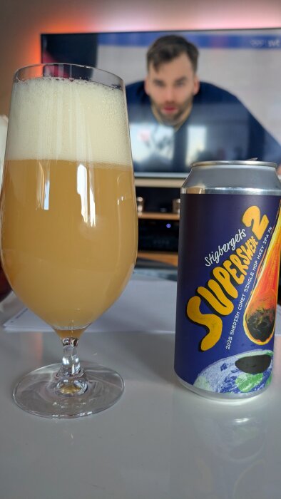 Glas med ljus öl bredvid en burk Stigbergets Superswede 2 öl. I bakgrunden syns en suddig TV-skärm.
