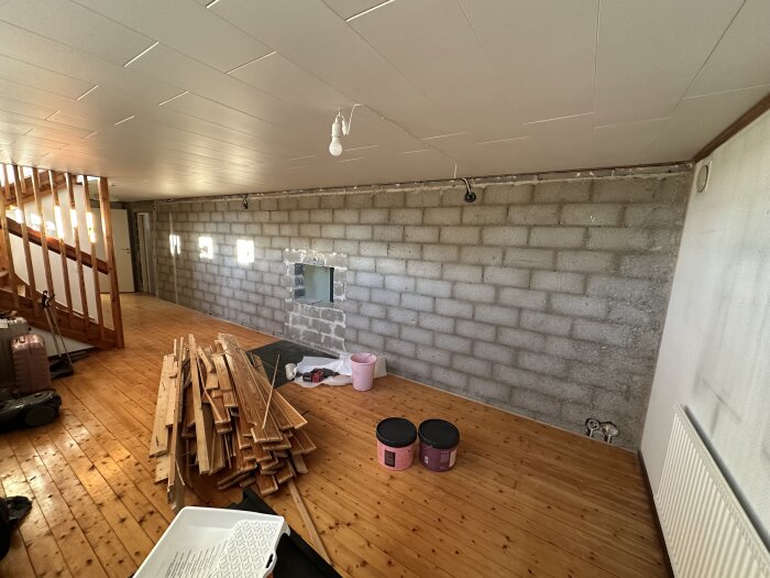 Renovering pågår i källare med putsarbete och ny kamininstallation. Staplar av träplank och målarhinkar på ett trägolv nära en gråstensvägg.