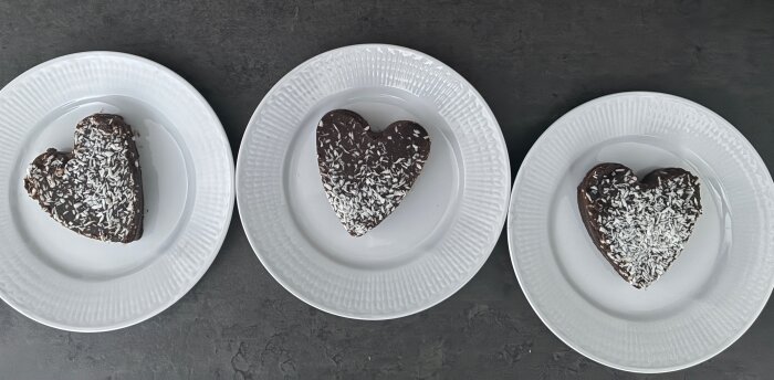 Tre hjärtformade chokladkakor med kokosflingor på vita tallrikar på en mörk yta.