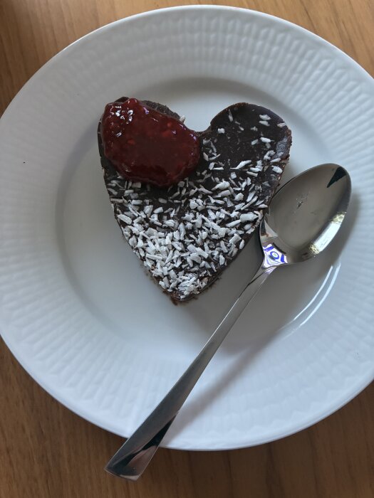Hjärtformat chokladbakverk med kokosflingor och sylt på ett vitt fat, bredvid en sked.