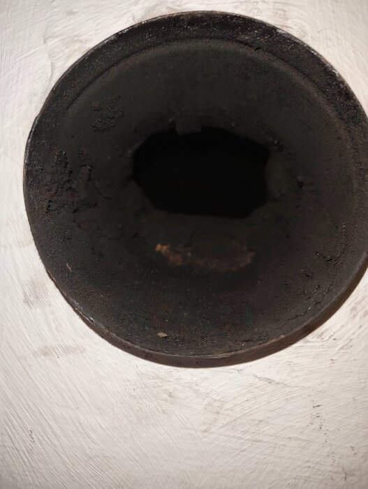 Rostigt metallrör synligt från sidan, inbäddat i vit vägg. Röröppningen är mörk och visar insidan av röret med en ojämn kant.