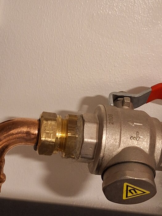 Närbild på en metallventil kopplad till en kopparrörledning. Ventilen har en röd spak och ett varningsmärke.