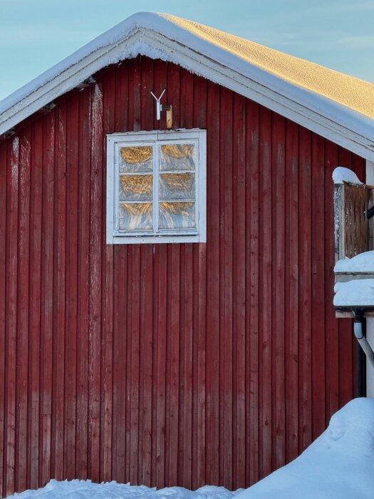 Röd träbyggnad med snötäckt tak och frostat fönster. Snö på marken, antenn monterad ovanför fönstret.