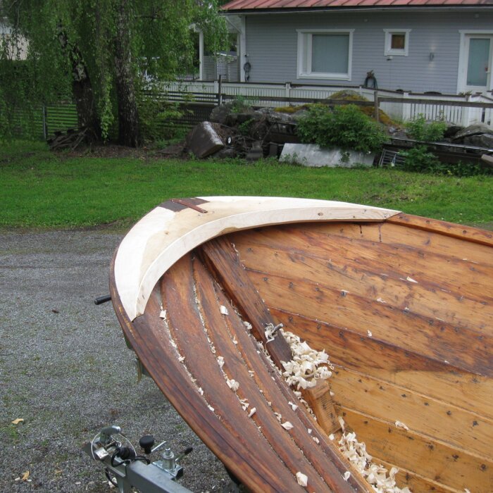 Träbåt under renovering med nytt virke och träspån, placerad på en gräsplan nära ett hus.