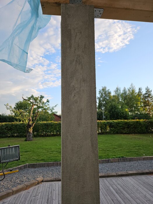 Putsad pelare på terrass med nät och hörnlister under ett trätak, omgiven av parkmiljö med träd och gräsmatta.