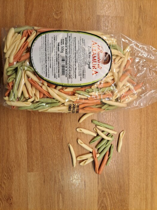 Förpackning med färgglad, torkad pasta på ett träbord, visar olika pastaformer i grönt, rött och beige.
