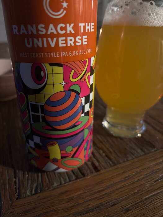En färgglad burk med "Ransack the Universe" West Coast IPA och ett glas öl på ett mörkt träbord.
