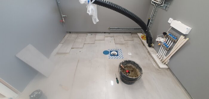 Rum under renovering med klinkerplattor, en hink med murbruk, verktyg, och rörinstallationer på väggarna.