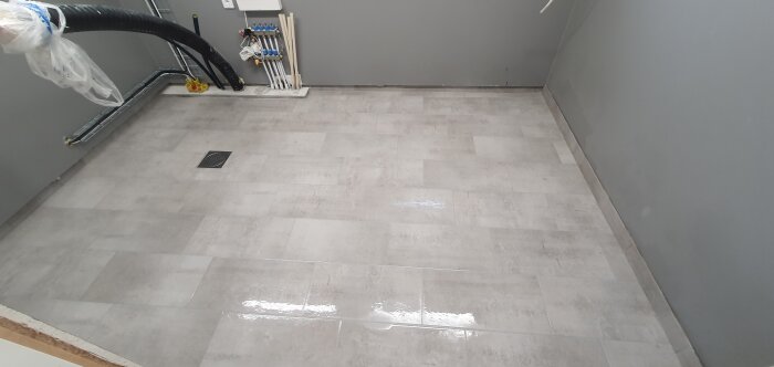 Grått klinkergolv i ett verktygsrum med avloppsbrunn och synliga rör längs väggen. Golvet är nyligen installerat.