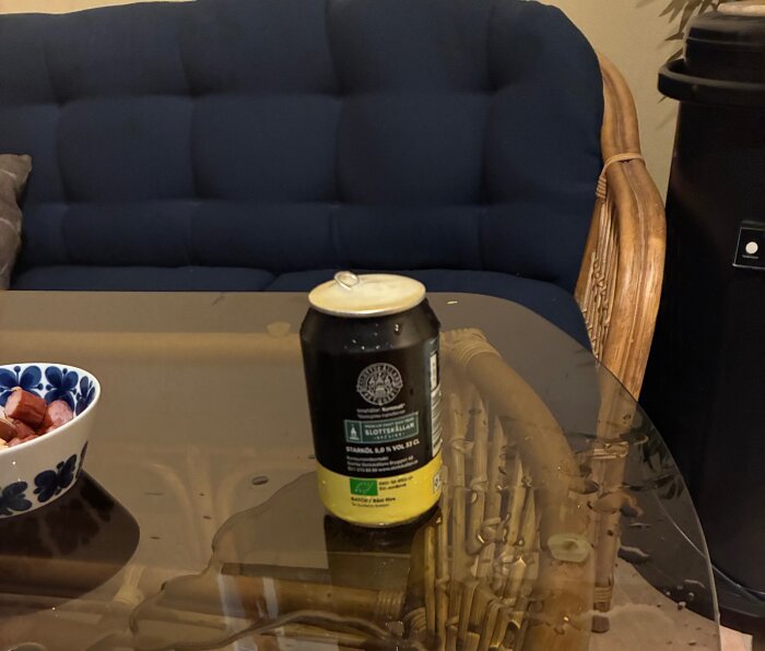 En öppnad ölburk på ett glasbord med spill bredvid, en blå soffa och en skål med korv.