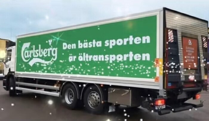 Lastbil med texten "Den bästa sporten är öltransporten" på sidan, logga synlig på en parkeringsplats.