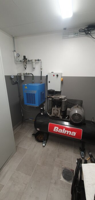 Kompressor och tork med rörledningar installerade på klinkergolv i verkstadsrum.