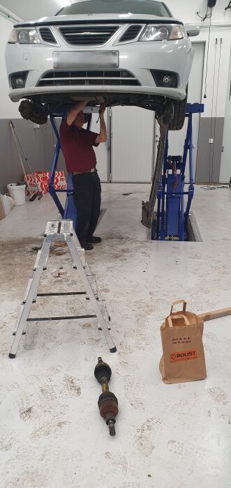 En person arbetar under en upphissad bil i ett garage, med verktyg och en papperspåse från Bolist på golvet.