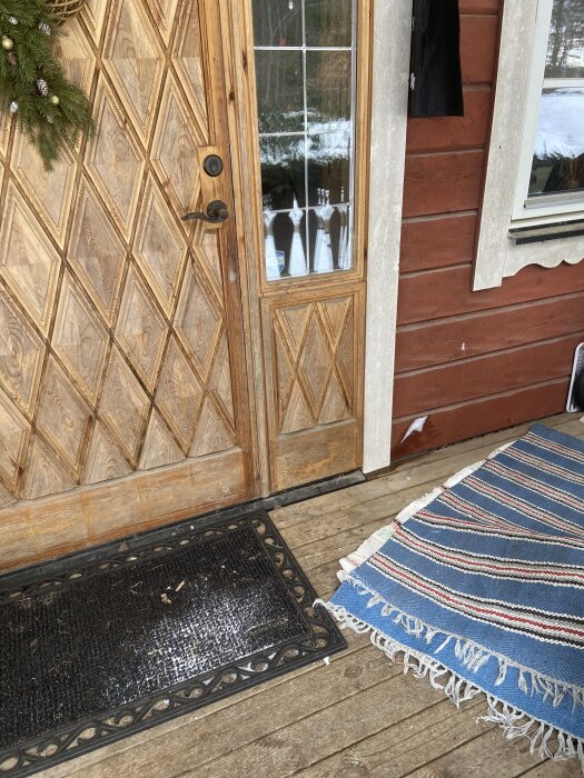 Ytterdörr och vägg med träpanel och glas vid ett hus. Det finns isbildning intill dörren och en randig matta samt dörrmatta syns på verandan.