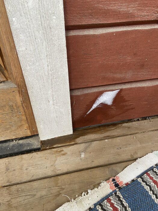Isbildning på en röd yttervägg nära en dörrkarm, ovanpå en trätröskel med tätning. En stickmatta syns på golvet.