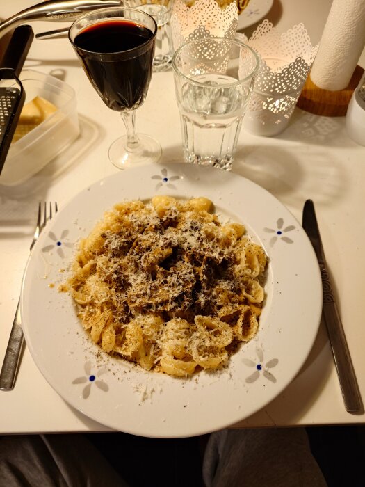 En tallrik lövbiffspasta toppad med riven ost och peppar, serverad på ett bord med rött vin och vatten.