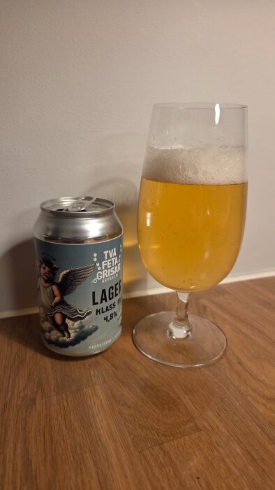 Ölglas med skummande lager och en ölburk med etikett från Två Feta Grisar bryggeri står på ett träbord.