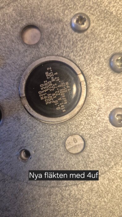 Närbild på ny fläkt med metallisk yta och märkning för 4uF kondensator, omgiven av skruvar.