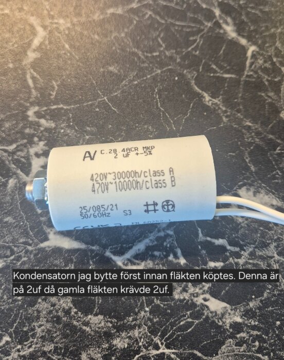 Kondensator på 2uF med text och tekniska specifikationer liggande på ett marmorerat underlag.