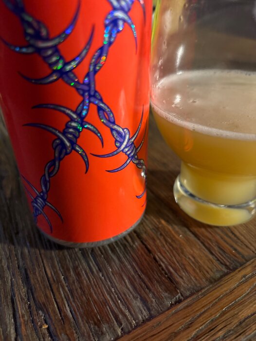 En röd burk med blått taggtrådsmönster bredvid ett glas med gul, skummande dryck på ett träbord.