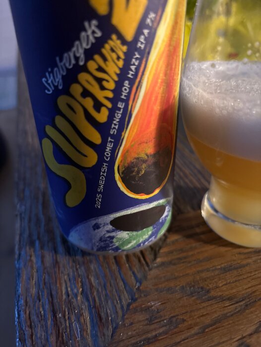 Burk med öl märkt "Stigbergets SUPERSWEDE" och ett glas med skummande öl, stående på ett träbord.