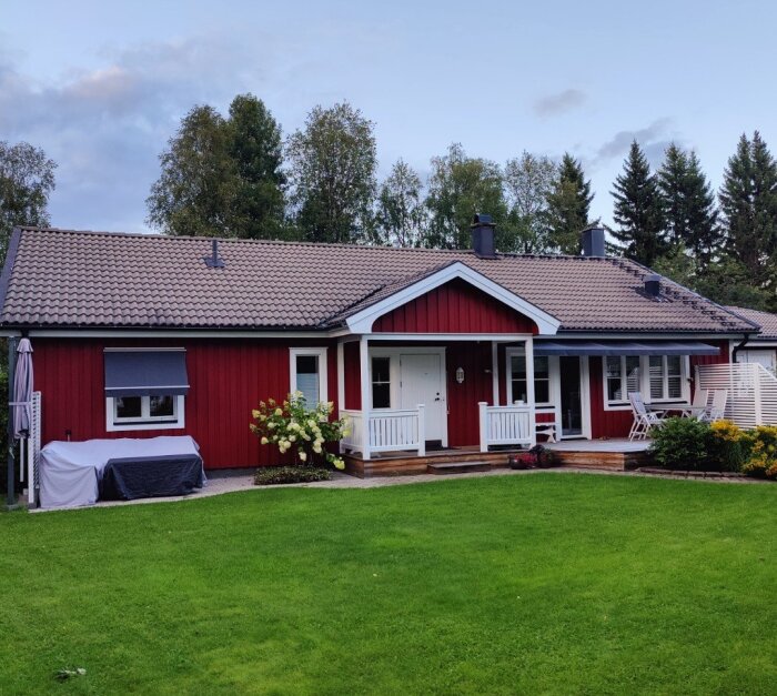 Rött enplanshus med betongtegelstak, omgiven av en grön trädgård med utemöbler på verandan.