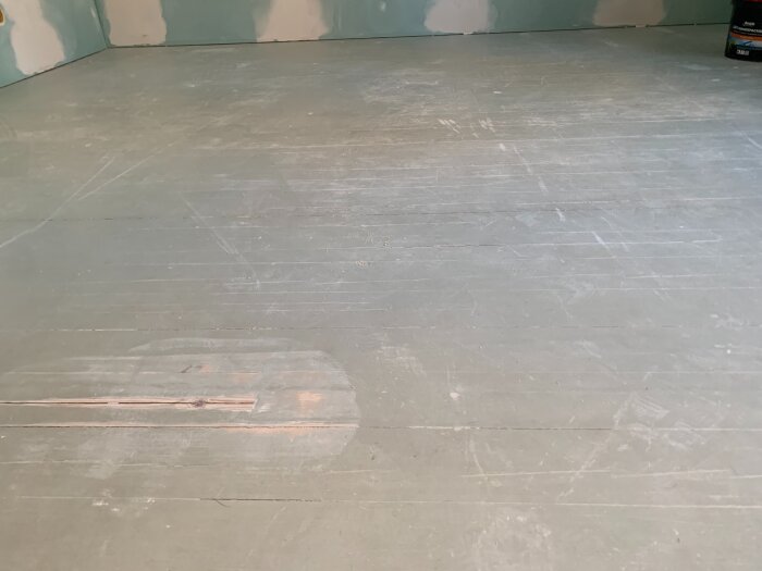 Slitet målat trägolv med sprickor, i behov av slipning och eventuell renovering. Bakgrunden visar ofärdiga väggar.