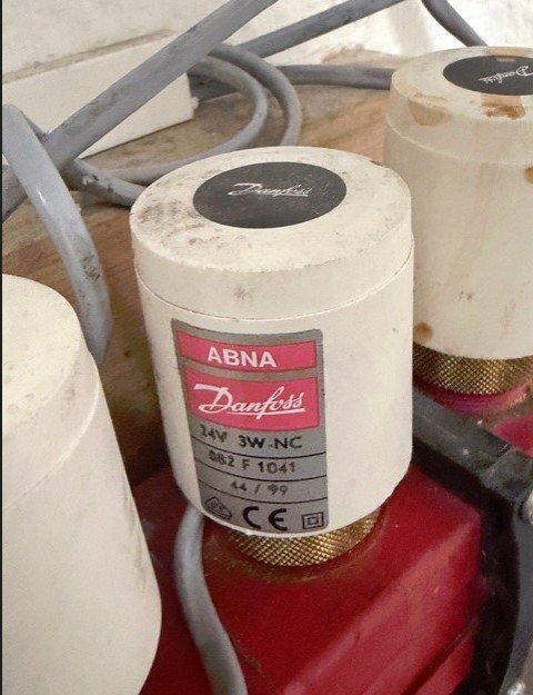 Termostat för golvvärme från Danfoss, märkt ABNA 24V 3W-NC, ovanpå röd bas med kablar i bakgrunden.