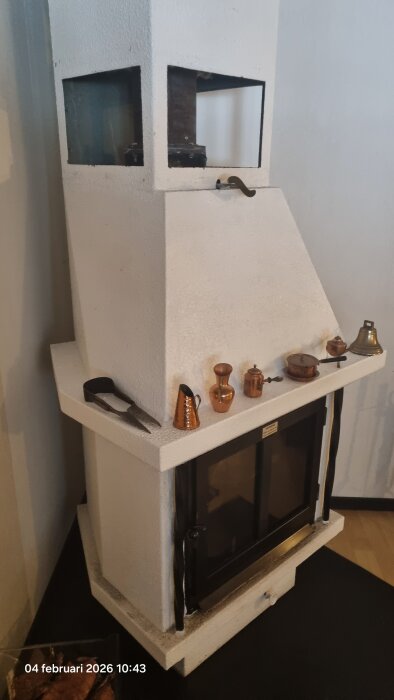 Vit Jormspis med en stor glaslucka och kopparföremål ovanpå, placerad i ett hörn av rummet.