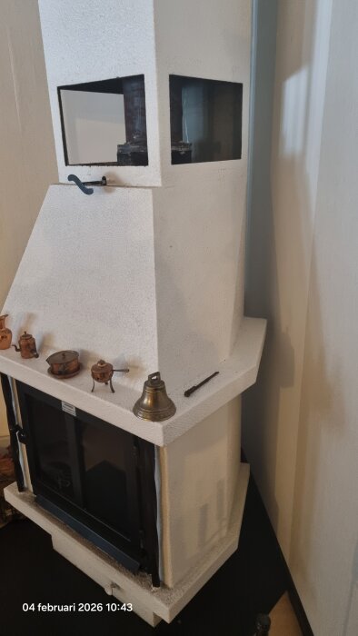 Vit Jormspis med två glasluckor, några kopparföremål ovanpå och en klocka, belägen i ett rum. Eldningsutrymmet är synligt upptill.