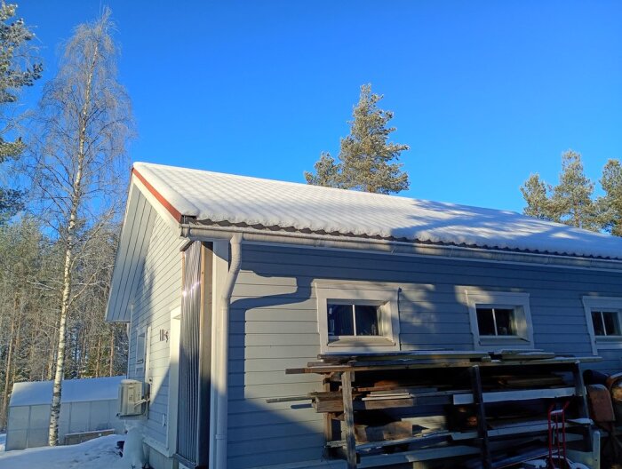 Blå himmel och ett snötäckt tak på grått garage med skuggor från träd. Träplankor staplade mot väggen, omgivna av skog och sol.