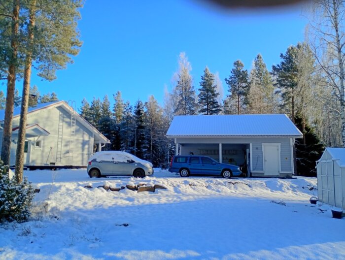 Snötäckt gårdsplan med garage och hus omgivna av träd, två bilar parkerade, vinterlandskap och klarblå himmel.