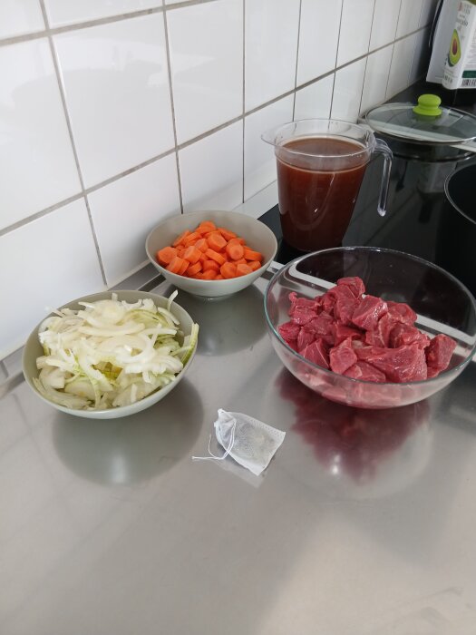 Skivad lök, morötter, köttbitar och en tesil på en bänk med en kanna brun vätska för att tillaga kalops.