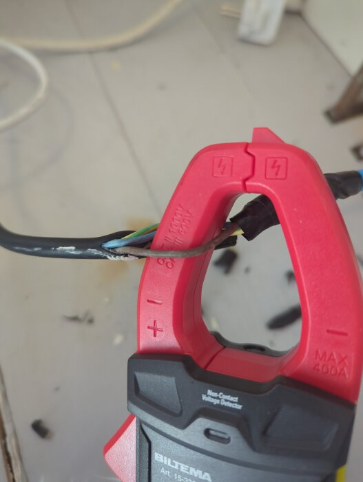 Multimeter klämd runt en elkabel för att mäta ström eller spänning, synlig på en byggarbetsplats med oregelbundet golv.