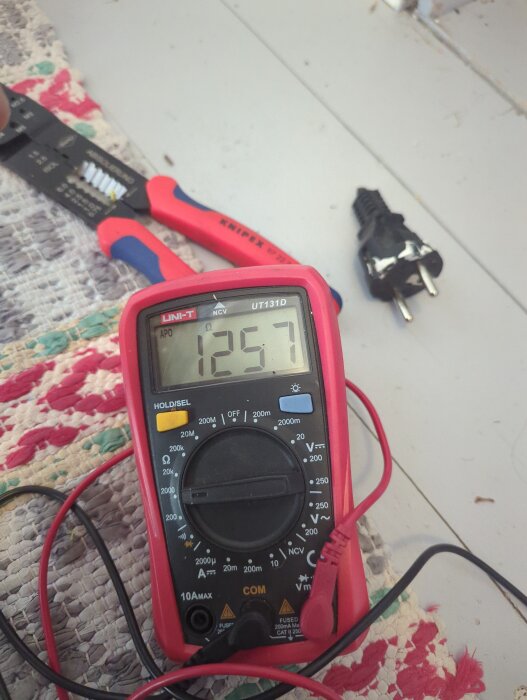 Multimeter visar 1257 ohm bredvid en svart stickpropp och en kabelskalare på en vävd matta.