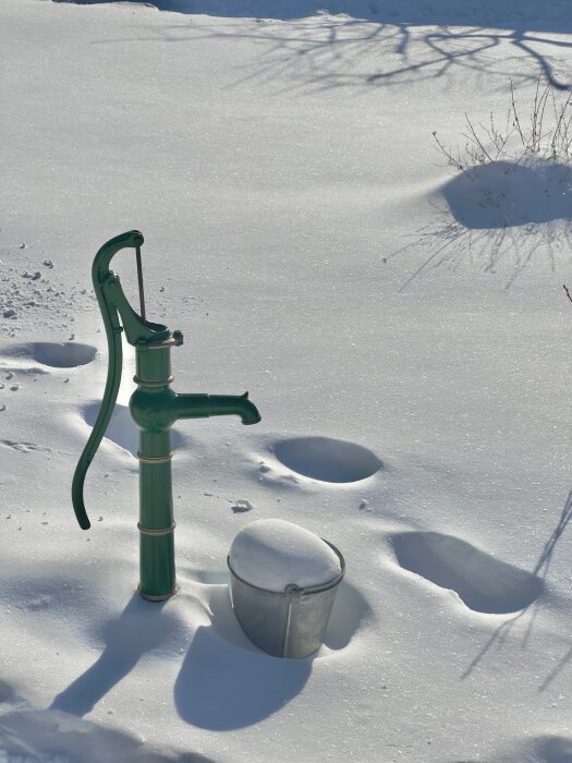 Grön vattenpump och en hink på snöig mark med skuggor från grenar.