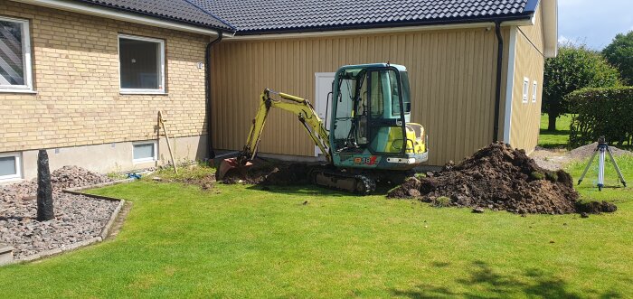 Minigrävare gräver vid sidan av ett garage, grön gräsmatta och en hög jord syns, projekt pågår att fixa området kring garaget.