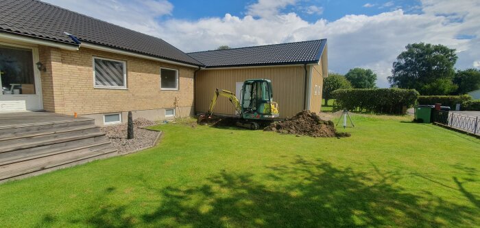 Minigrävare vid garage, utgrävd jordhög, hus med trappa, grön gräsmatta, byggprojekt pågår.